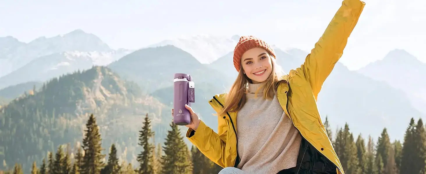 A happy woman in a yellow jacket holding a PureSip Thermal Bottle in the mountains, enjoying the outdoors.