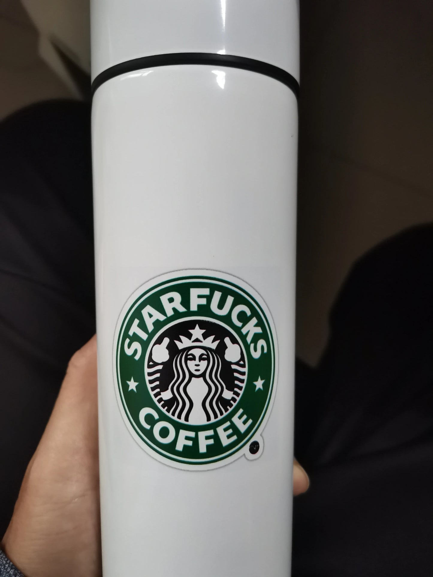 Close-up of a white travel mug featuring a humorous Starbucks parody logo with the text 'STARFUCKS COFFEE'.